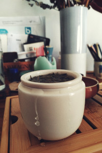 A vertical colour photograph of a tea cup resting on a bamboo tray. The tea cup is full of water and long light green leaves brewing. On the ceramic, a trail of water has escaped from the cup.