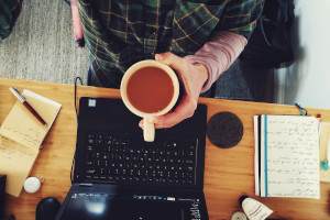 A top down horizontal colour photograph of my hand holding a mug of tea over a desk with a laptop, paper, a pen, and various desk paraphernalia.