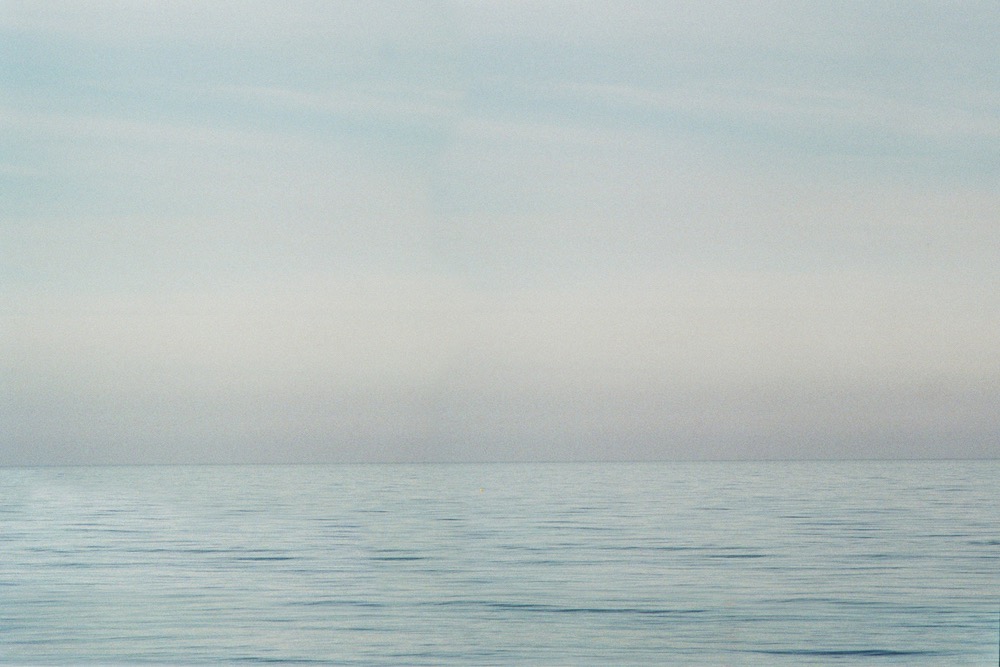 A soft colour photograph of the seaside. The top two third contains a featureless sky, the bottom third contains the sea and gentle ripples on its surface.