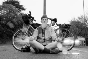 A black and white photograph of me (a white woman with dark hair) sitting cross legged on tarmac with my eyes closed. Behind me is a bicycle. On either side of me blurred cars are driving past. Drops of rain are visible on the camera lens.