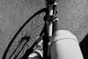 A horizontal black and white photograph of my legs pedalling. The front wheel cast a shadow on the tarmac. I am wearing a light coloured short and light coloured trainers.