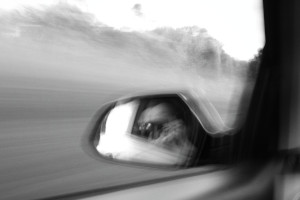A horizontal black and white photograph of me creating a photograph in the rear-view mirror of a car. The car is in motion so the image is entirely blurred with nothing well defined.