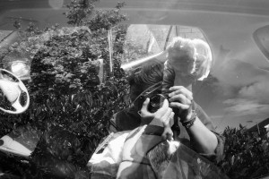A horizontal black and white photograph of me (a while person with short black hair and rolled up sleeves) reflected in a car window. The scene shows the inside of the car as well as bushes from the outside and a person with short dark hair glowing in the sun bent over a seat from an open car door.