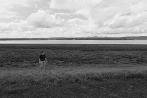 A horizontal black and white photograph of me standing in a layered farmed flat landscape. I am a white person with short dark hair. I am standing in a field, wearing a light coloured short and dark coloured hoodie (with rolled-up sleeves. Beyond me is a river, a thin strip of land, and clouds above.