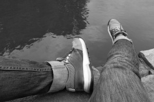 A horizontal black and white photograph of a close-up of my legs from the knee down resting on stones by water's edge. I am wearing dark jeans and trainers.