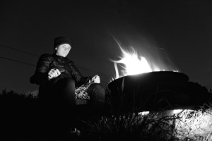 A horizontal black and white photograph of me by a campfire at night. I am a white person wearing a dark outfit from leggings to jacket to beanie.
