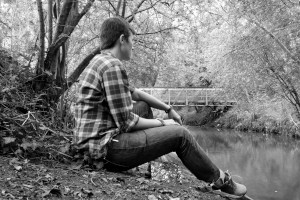 A black and white horizontal photograph of me (a white woman with short dark hair, face mostly turned away from the viewer. I am wearing dark jeans, trainers, and a checkered shirt with rolled up sleeves) sitting by a river with a footbridge in the background.