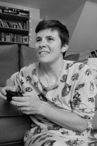A vertical black and white photograph of me (a white woman with short dark hair) in my pyjama, playing a video game. I am sitting by a grey sofa, holding a black Nintendo Switch controller.