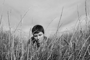 A horizontal black and white self-portrait. I am a white woman with short dark hair, lying in tall grass. Only my shoulders and head are visible through a curtain of grass. Above, the sky is full of clouds.