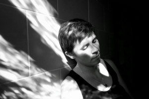 A horizontal black and white self-portrait bathed in dappled light. I am leaning against a tiled wall, eyes closed, following the trail of the light. I am a white woman with short dark hair wearing a dark tank top.