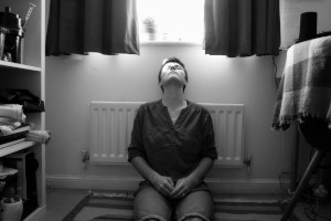 A horizontal black and white self-portrait of me sitting against a radiator by a wall under a window. Light streams from the window and fall on one of my eyes. I am a white person with short dark hair wearing a light coloured short and a dark coloured shirt. My head is tilted back towards the light and my hands are together resting on my legs.