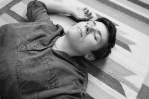 A horizontal black and white photograph me from the chest up - a white person with short black hair, eyes closed, a dark shirt with rolled up sleeves - lying diagonally on a carpet.