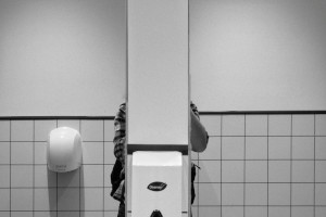 A horizontal black and white photograph of me in a public bathroom. I am creating the photography using my reflection in two mirrors, however I have placed my body so that most of me is in the gap between the mirror. Only part of my arms and legs are visible, the rest if a white wall.