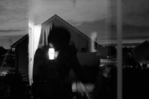 A horizontal black and white photograph of the outline of my hunched body reflected in a window. Outside the window is a brick house in suburbia.
