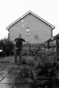 A vertical black and white photograph of me standing in a suburban garden in the rain. I am a white woman with short dark hair. I am wearing a dark t-shirt and dark part of shorts. I am barefoot. I am in the act of jumping, my body blurred in the air.
