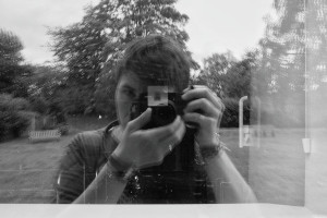 A horizontal black and white photograph of me (a white woman with short dark hair) holding a camera in front of my face. My image is reflected in a window. Some of the building inside can be glimpse but mostly a garden us reflected being me.