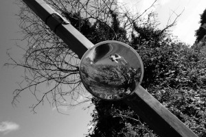 A horizontal black and white photograph of me (a white woman) wearing a helmet. My full body is reflected in a round street mirror.