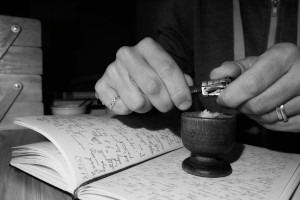 A horizontal black and white photograph of my hands sharpening a pencil in a wooden eggcup resting atop a notebook.