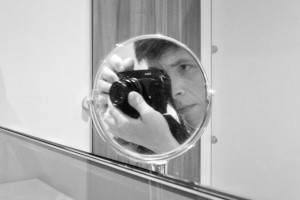 A horizontal self-portrait in a shaving mirror reflected in a wider wall long mirror. I am a white person with short black hair. I am only reflected in the shaving mirror, with part of my face visible and some of my hand holding a black camera.