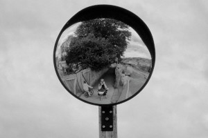 A square black and white photograph me of (a white woman) reflected in a street mirror with a camera over my face. I am straddling my bicycle.