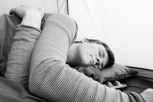 A horizontal black and white photograph of me (a white woman with short dark hair) sleeping in a tent with arms crossed. I am visible from the chest up.