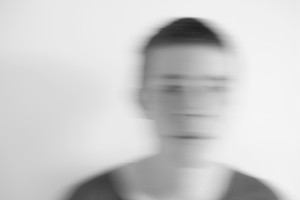 A horizontal black and white photograph of my face (a white woman with cropped black hair) blurred by movement.