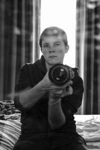 A vertical black and white photograph of my face (a white woman with short black hair). I am sitting on a bed holding a camera in front of my chest.