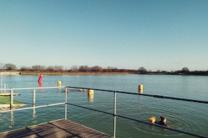 A horizontal colour photograph of a still lake under a winter sunny blue sky. A few folks are swimming. Yellow and red buoys float atop the water. In the distance bare trees lie the lake. In the foreground there is a fence with it's shadow on brown decking.