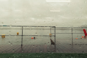 A horizontal colour photograph of a lake with people swimming in it seen through a window splattered with rain drops.