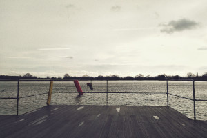 A horizontal colour photograph of a lake with choppy water. In the foreground is some wood like decking and metal barriers.